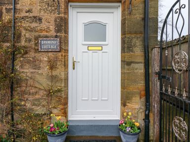 A white door with a mail slot and gold handle flanked by flower pots and a stone wall with a Jasmine Cottage sign