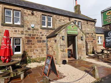 Exterior of a stone building with signs for The Coach Inn bar restaurant and outdoor seating with red umbrellas at Jasmine Cottage in Lesbury near Alnmouth