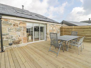 An outdoor wooden deck with a metal table and chairs next to a stone building with glass sliding doors at Horseshoe Cottage in Abersoch