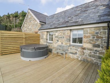 An outdoor wooden deck with a covered hot tub and a metal bench beside a stone cottage at Horseshoe Cottage in Abersoch