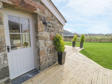 A stone building with a white door and outdoor plants on a wooden deck overlooking a grassy area at Horseshoe Cottage in Abersoch