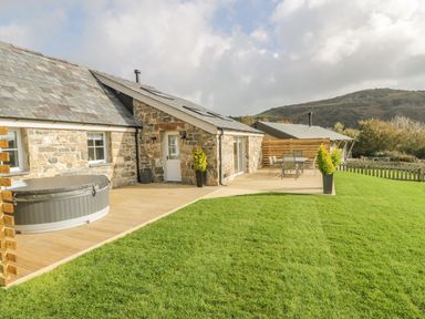 A stone cottage with a wooden deck featuring a hot tub and outdoor dining set surrounded by grass at Horseshoe Cottage in Abersoch