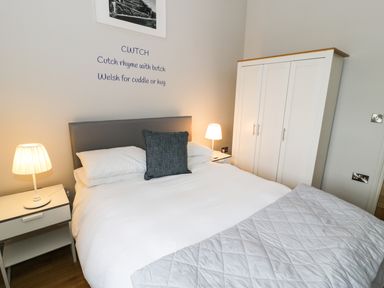 A bedroom with a bed with white linens a gray pillow two bedside tables with lamps and a white wardrobe at Horseshoe Cottage in Abersoch