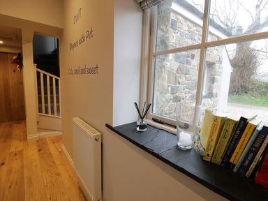 A hallway with wooden floor and staircase a window ledge with books a glass with ice and a reed diffuser at Horseshoe Cottage in Abersoch