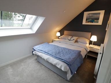 A bedroom with a bed a blue quilt two bedside tables with lamps a skylight window and a framed picture at Horseshoe Cottage in Abersoch