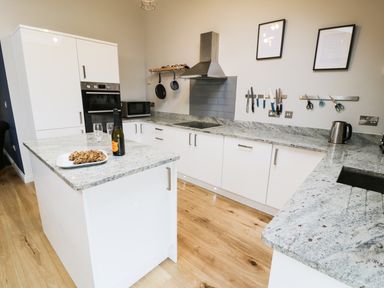 A kitchen with white cabinets granite countertops a built in oven a microwave hanging pans a kettle knives and scissors on magnetic strips and a bottle of wine with two glasses and cookies on the countertop at Horseshoe Cottage in Abersoch
