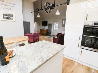 A kitchen with a granite countertop an oven a wooden table and chairs and a living area with armchairs and a tv at Horseshoe Cottage in Abersoch