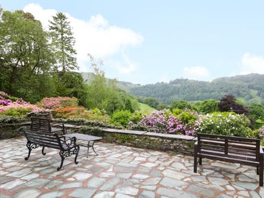 An outdoor patio area with stone flooring wooden benches a metal table and green hills in the background at Ramblers Roost in Grasmere