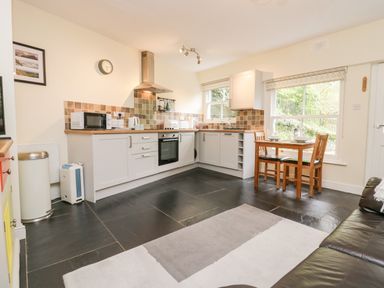 A kitchen with white cabinets and tiled backsplash a wooden dining table with two chairs and a black leather sofa at Ramblers Roost in Grasmere