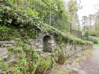 A stone wall with plants growing on it next to a paved path and metal railing with trees in the background at Ramblers Roost in Grasmere