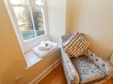 A corner with a window seat holding folded towels next to a velvet armchair with a checkered cushion at East Farmhouse Cottage in Humshaugh near Hexham