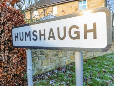 A roadside sign reading Humshaugh with a stone building and hedges behind it at East Farmhouse Cottage in Humshaugh near Hexham