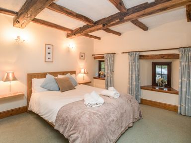 A bedroom with a double bed pillows rolled towels bedside tables lamps wooden beams and small windows with curtains at Riven Oak in Lyth Valley near Levens
