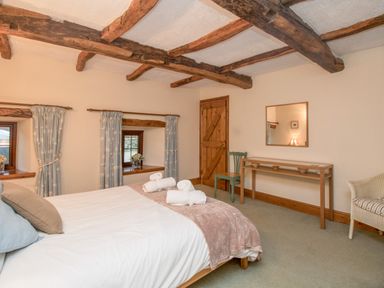 A bedroom with exposed wooden beams bed with rolled towels two windows with curtains a chair wooden door and a table with a mirror at Riven Oak in Lyth Valley near Levens