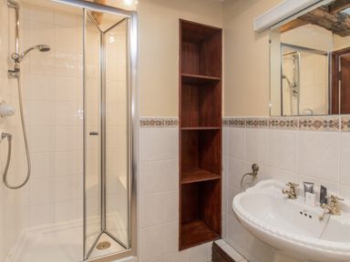 A bathroom with a corner shower enclosure a wooden open shelf a white sink with faucets and toiletries at Riven Oak in Lyth Valley near Levens