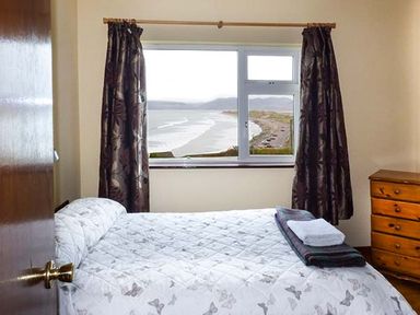 A bedroom with a bed covered in a butterfly patterned quilt and folded towels on top next to a window with curtains overlooking a beach at Rossbeigh Beach Cottage No 6 in Rossbeigh near Glenbeigh County Kerry