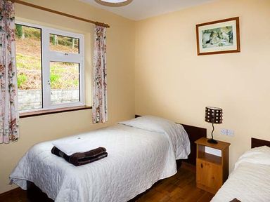 A bedroom with two single beds white bedding a window with floral curtains and a wooden nightstand with lamp at Rossbeigh Beach Cottage No 6 Rossbeigh near Glenbeigh County Kerry