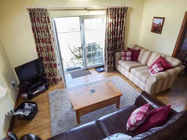 A living room with a wooden coffee table a beige striped sofa with red pillows a dark brown leather sofa with patterned pillows a TV on a stand and glass doors leading outside at Y Barcud in Holyhead
