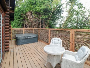 An outdoor wooden deck with a hot tub and two white wicker chairs with a round table at Carlton Lodge 1 in Stokesley