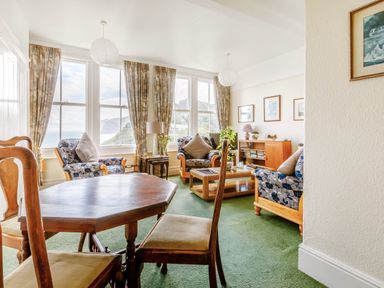 A living room with wooden chairs and a table near large windows with curtains at Clooneavin Apartment 3 in Lynmouth near Lynton
