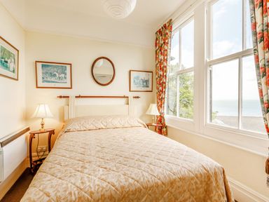 A bedroom with a bed two bedside tables with lamps framed pictures a round mirror and large windows with floral curtains at Clooneavin Apartment 3 Lynmouth near Lynton
