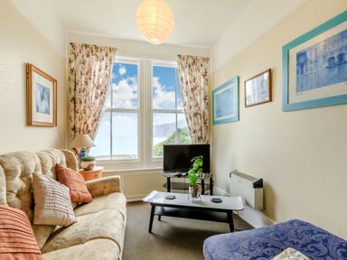 A small living room with a beige sofa and patterned blue chair facing a TV on a stand by a window with floral curtains at Clooneavin Apartment 5 in Lynmouth near Lynton