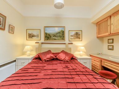 A bedroom with a red bedspread and pillows two bedside tables with lamps paintings on the wall and wooden cupboards with a countertop at Clooneavin Apartment 5 in Lynmouth near Lynton