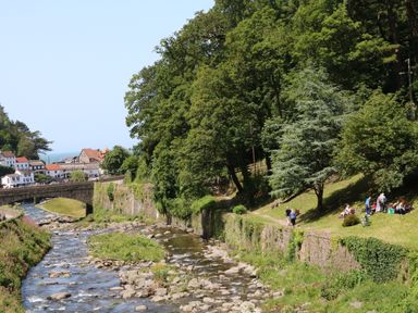 A river with rocks flowing under a stone bridge beside a grassy area with people sitting and walking near trees and houses in the background at Clooneavin Apartment 8 in Lynmouth near Lynton