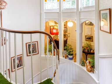 A curved staircase with white railings and green carpet beside windows with plants and framed pictures on the walls at Clooneavin Apartment 8 in Lynmouth near Lynton
