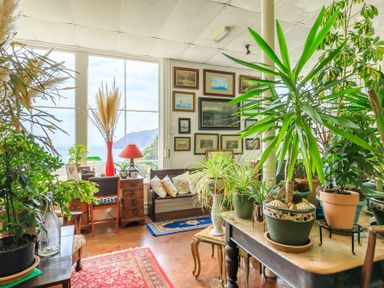 A room with large windows overlooking a mountain and sea with multiple plants wooden furniture and framed pictures at Clooneavin Apartment 8 Lynmouth near Lynton
