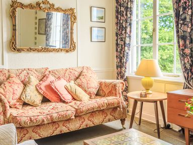 A living room with a patterned sofa cushions a wooden side table with lamp and a window with floral curtains at Clooneavin Apartment 8 Lynmouth near Lynton