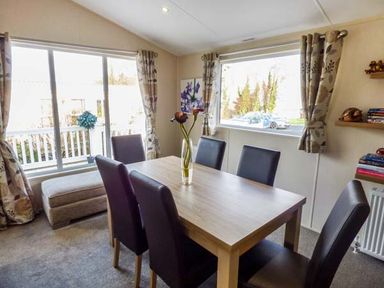 A dining room with a wooden table six chairs and windows with curtains at Clearview Lodge in Borth