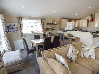 A living room with a beige sofa and floral cushions a dining table with six chairs and a kitchen with cabinets at Clearview Lodge in Borth