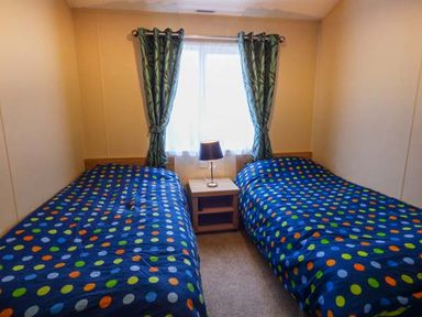 A bedroom with two single beds with blue polka dot bedding a nightstand and a lamp under a window with green curtains at Clearview Lodge in Borth