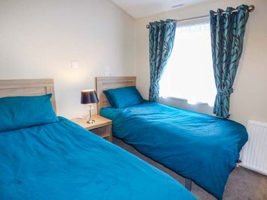 A bedroom with two single beds blue bedding a bedside table with lamp and window with curtains at Clearview Lodge in Borth