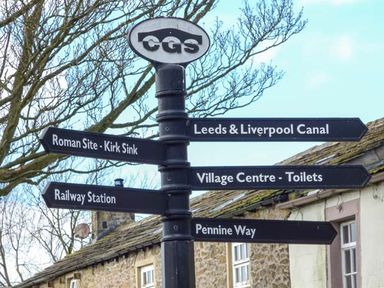 A black metal signpost with directional signs for Roman Site Kirk Sink Railway Station Leeds Liverpool Canal Village Centre Toilets and Pennine Way in a village setting