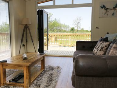 A living room with a wooden coffee table a gray sofa and a floor lamp next to an open door leading to a wooden deck at Squirrel Lodge in Gargrave near Skipton