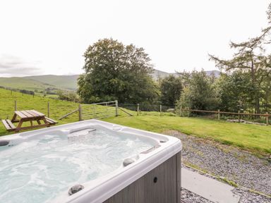 An outdoor hot tub on a patio with a wooden picnic table and a fenced grassy area with trees and hills at Bryndan in Machynlleth