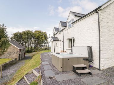 An outdoor hot tub beside a white stone house with a gravel path and steps leading to a stone building with red-framed windows at Bryndan in Machynlleth