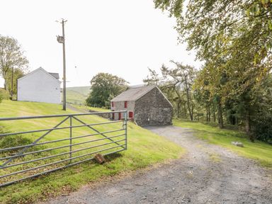 A gravel driveway leading to a stone barn with red windows near a white house and trees at Bryndan in Machynlleth