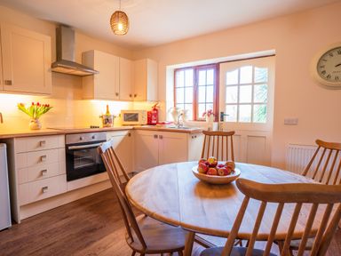 A kitchen with round wooden table and chairs a bowl of apples oven microwave flowers and a door with window at Nyth Y Wennol in Abersoch