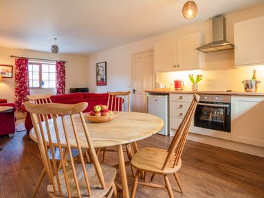 A kitchen and dining area with wooden table and chairs a bowl of apples white cabinets and a living room with red sofas and curtains at Nyth Y Wennol in Abersoch