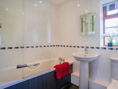 A bathroom with a bathtub, pedestal sink, toilet, window with blinds, and a red bath mat at Nyth Y Wennol in Abersoch