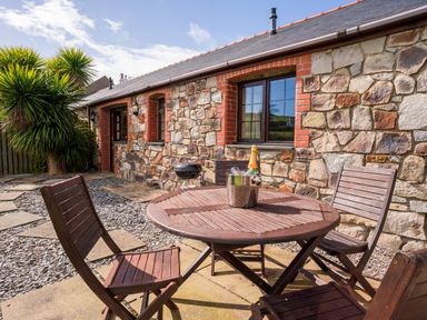 An outdoor patio area with a round wooden table and four chairs and a stone building in the background at Nyth Y Wennol in Abersoch