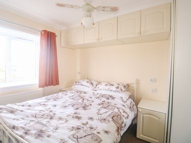A bedroom with a bed with floral bedding two bedside cabinets overhead cabinets red curtain and a ceiling fan at 11 Pendarves in St Merryn near Padstow