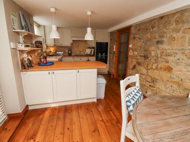 A kitchen with white cabinets a wooden countertop pendant lights a round wooden table a chair with a cushion and a stone wall at Edendale in Longframlington near Alnwick