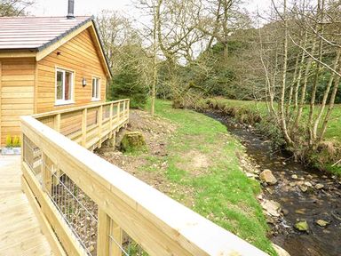A wooden cabin with a railing deck next to a small stream and trees at Park Brook Retreat in Scorton