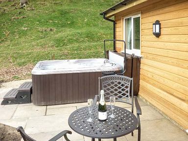 An outdoor patio with a hot tub next to a wooden building and a table with two chairs a bottle and two glasses at Park Brook Retreat in Scorton