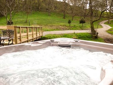 A bubbling outdoor hot tub with a wooden bridge and green trees in the background at Park Brook Retreat in Scorton
