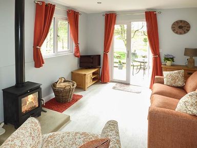 A living room with a wood-burning stove a wicker basket with firewood a television on a wooden stand orange curtains a wooden sideboard a table lamp and an orange sofa at Park Brook Retreat in Scorton